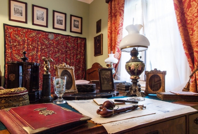 Zdrewniane biurko z umieszczonymi na blacie dokumentami, fajką i okularami. Na biurku stoi stara lampa naftowa. Na ścianie obrazy i kilim.