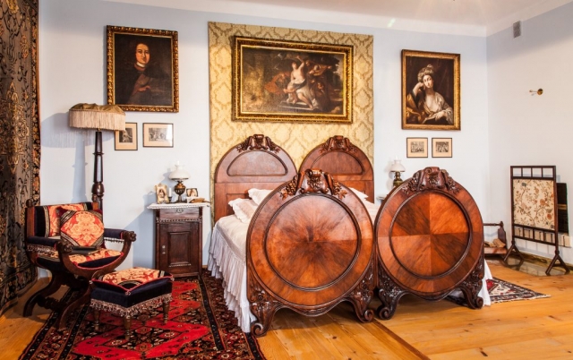Wnętrze dworku. Duże drewniane, zdobione łóżko, obok fotel i podnóżek. Na ścianach portrety w złotych ramach.