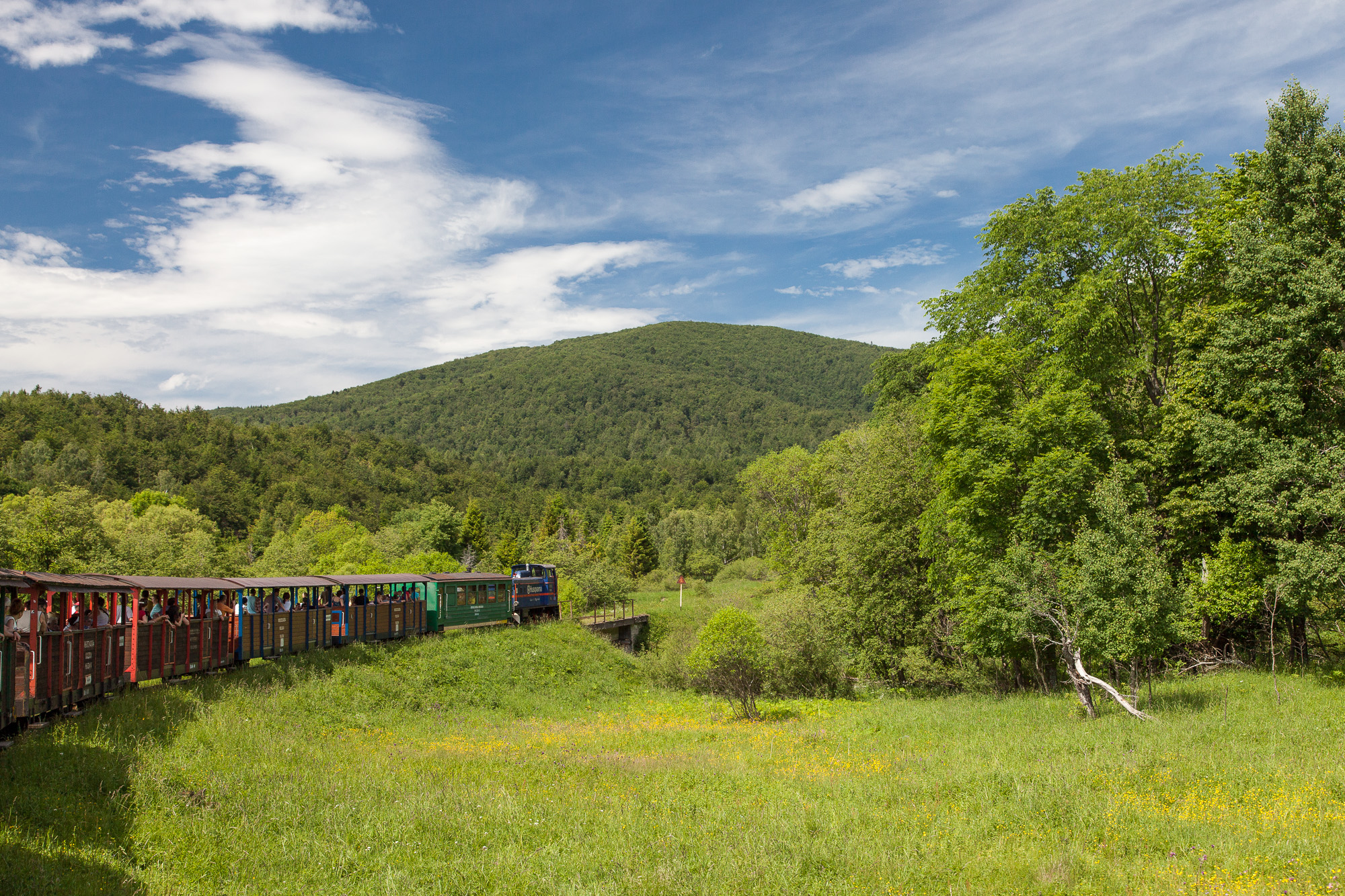 Letni dzień. Pociąg z wagonami jedzie po torach. Widać ludzi w otwartych wagonikach. W tle pokryta trzewami góra.