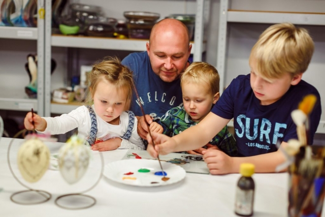 Trójka dzieci maluje farbkami na szkle. Pomaga im ojciec.