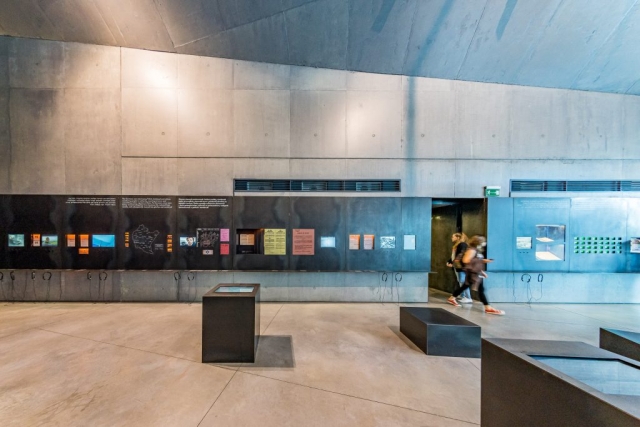 Wnętrze muzeum. Na ścianie zamontowane multimedialne ekrany z ekspozycją, obok zawieszone plakaty i obwieszczenia z czasów II wojny światowej.