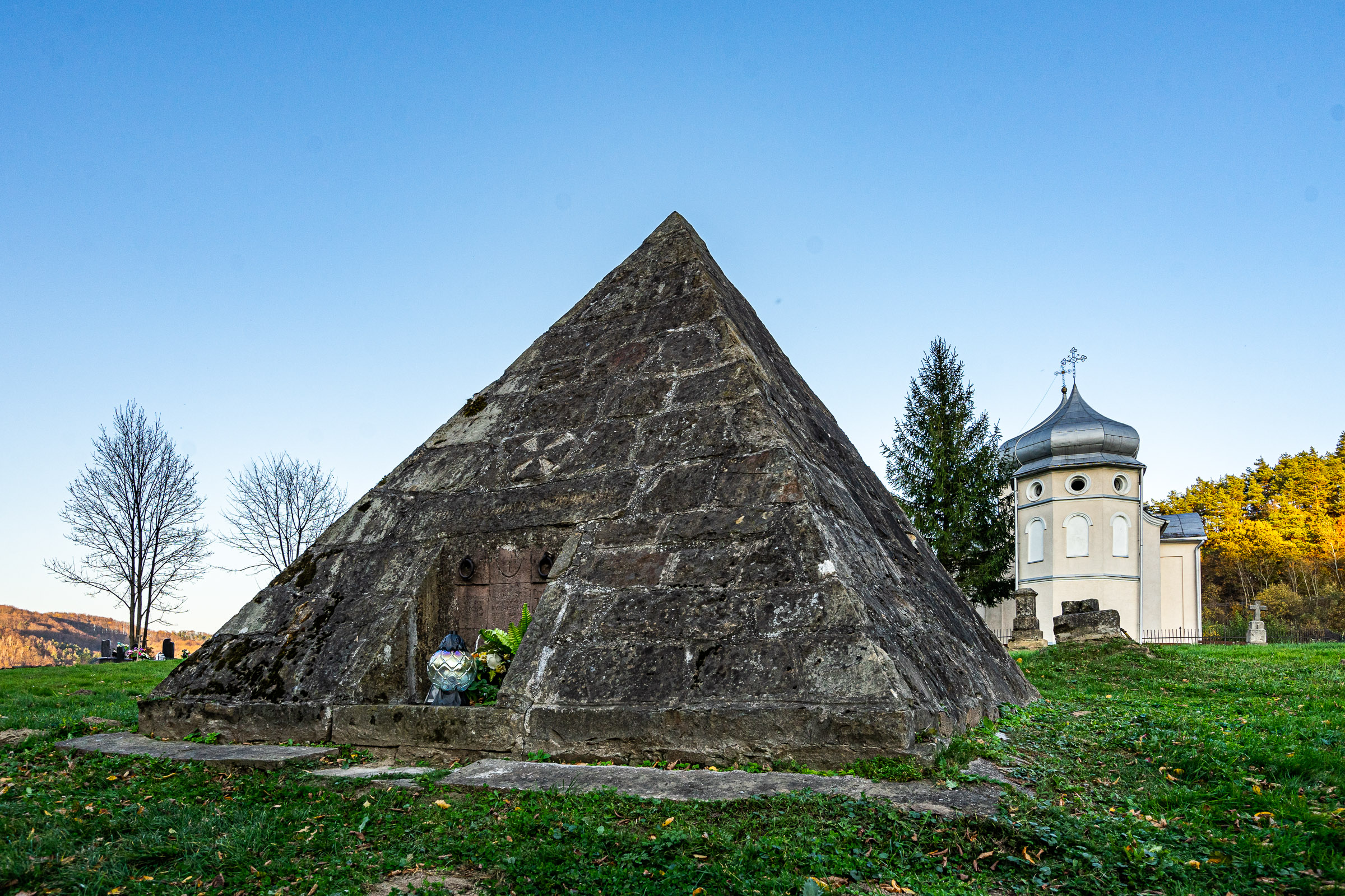 Kamienny grobowiec w kształcie piramidy. W tle budynek murowanej cerkwi.