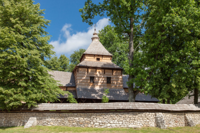 Drewniana cerkiew otoczona kamiennym murem pośród drzew.