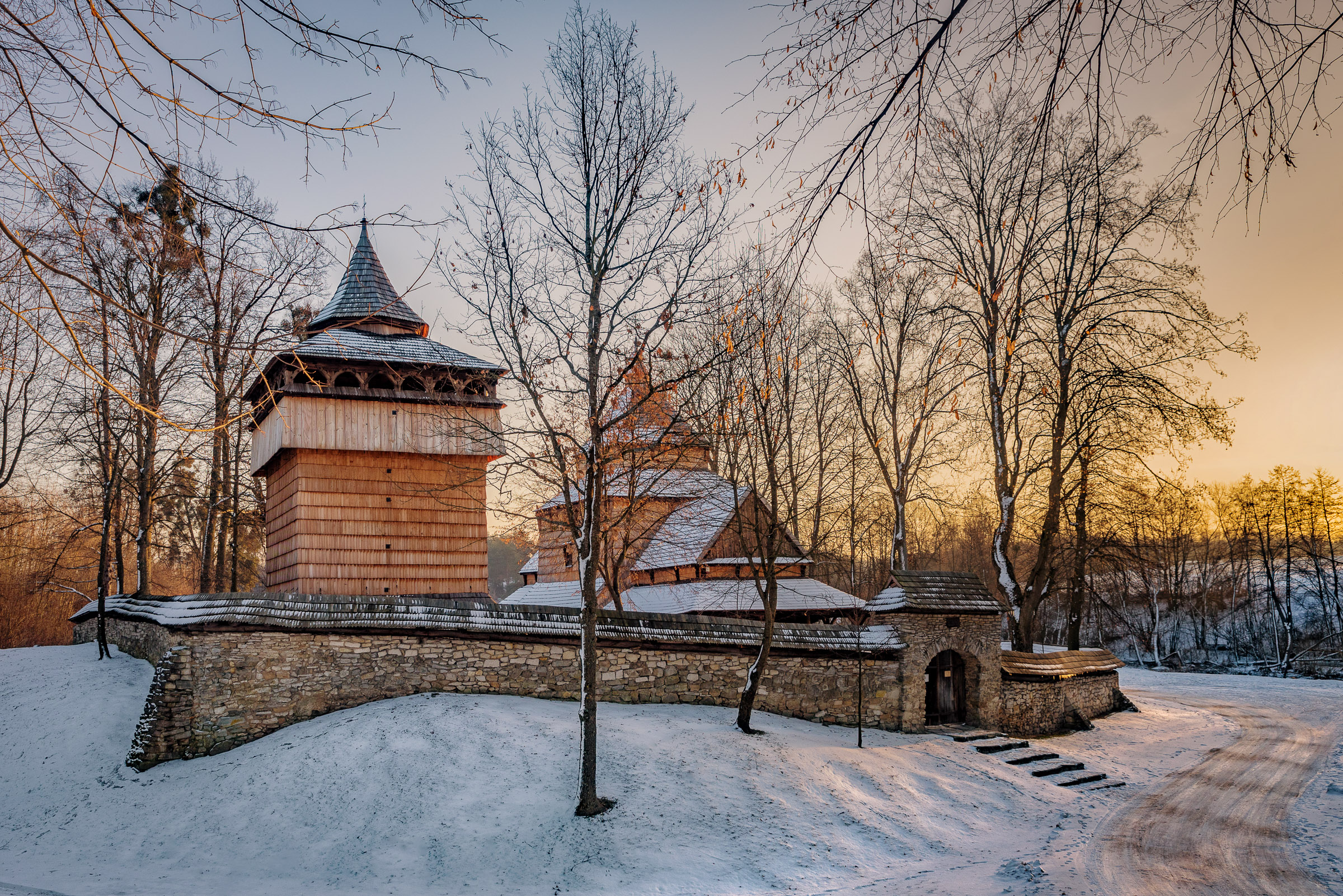 Zimowy zachód słońca. Drewniana cerkiew otoczona kamiennym murem pośród drzew.