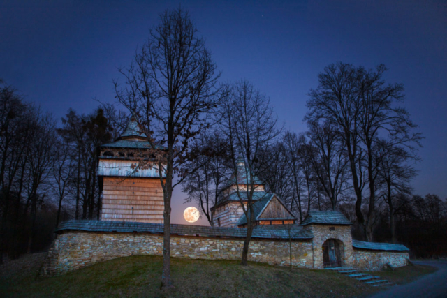 Noc. Księżyc w pełni między drewnianą dzwonnicą a cerkwią otoczoną kamiennym murem.