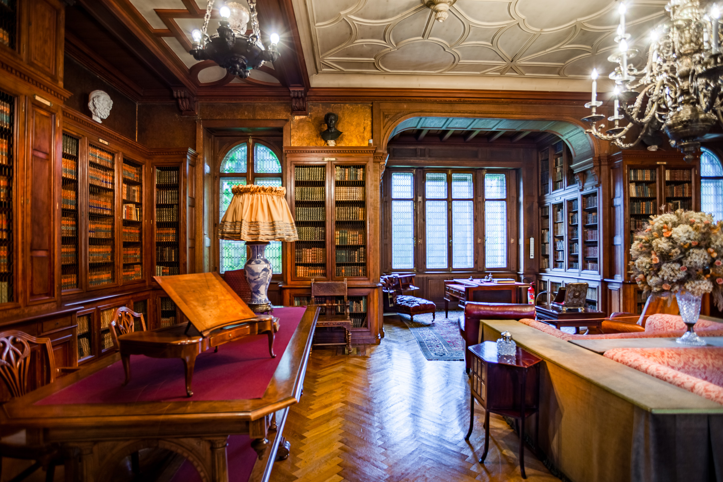 Eleganckie wnętrze w stylu angielskim. Na środuku sofa i skórzane fotele. Przy ścianach drewniane, zdobione regały z książkami. Na suficie zawieszony wielki żyrandol.