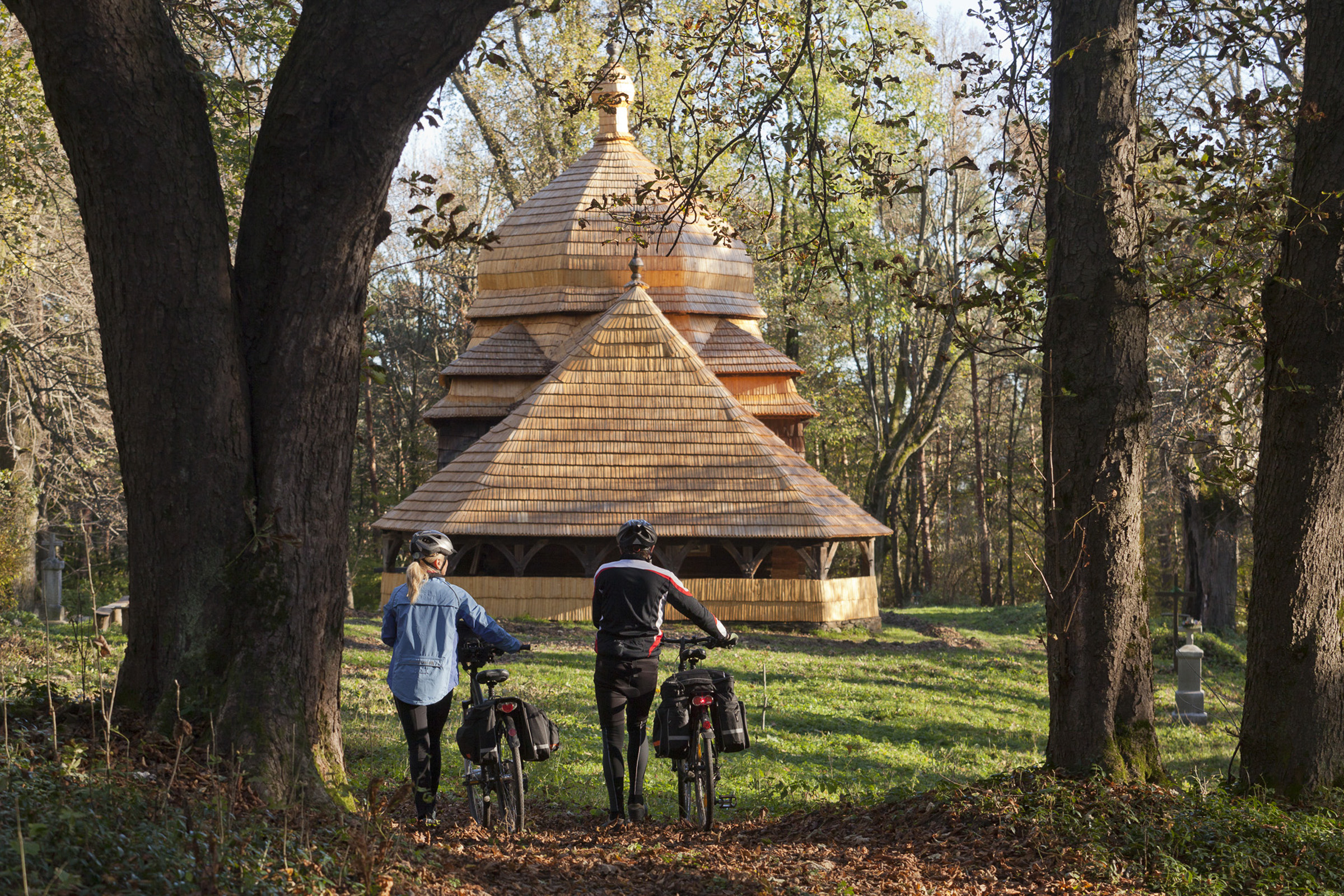 Dwoje rowerzystów prowadzących rowery leśną drogą w kierunku drewnianej cerkwi.