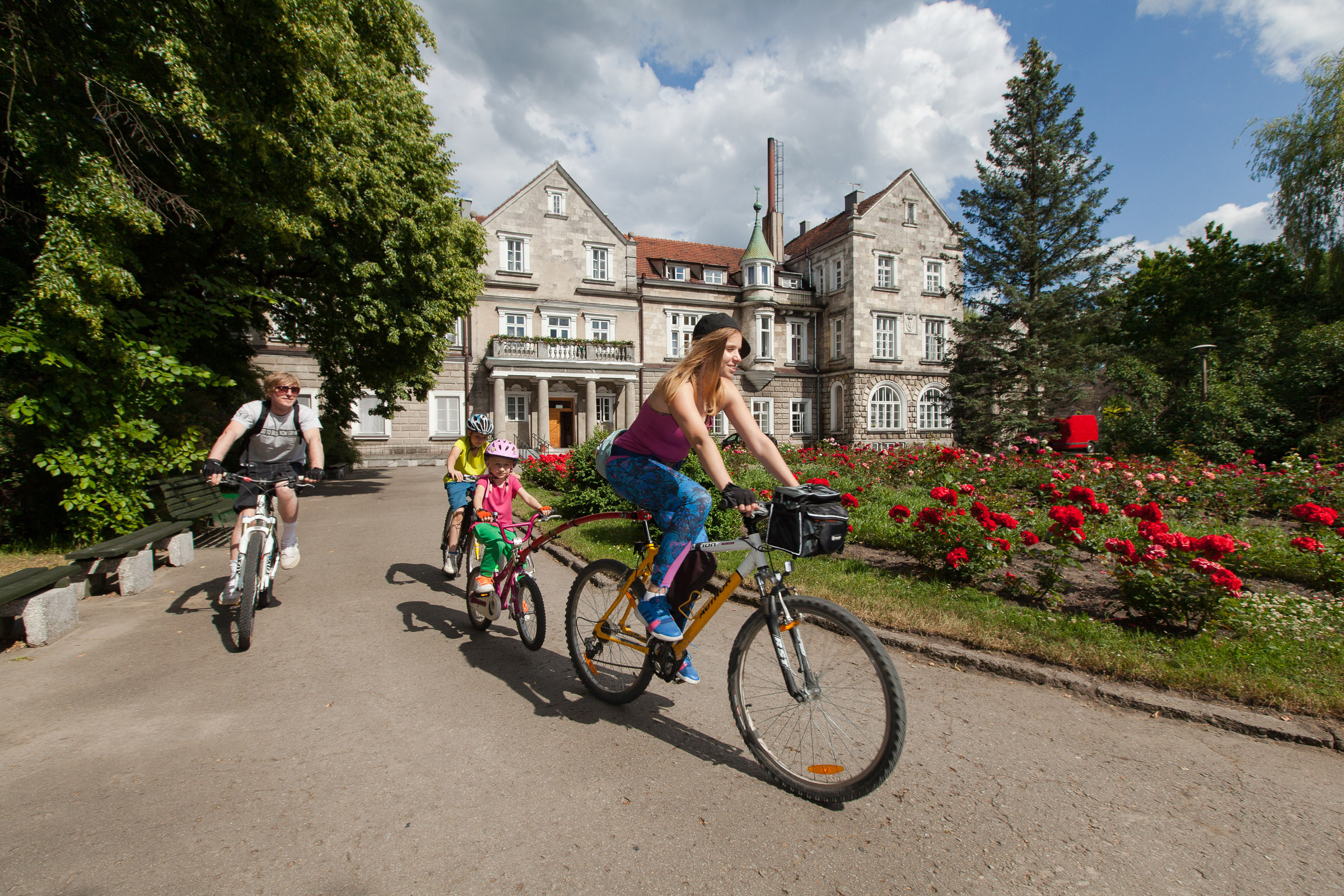 Grupa rowerzystów jadąca po drodze przed starym kilkupiętrowym budynkiem sanatorium.