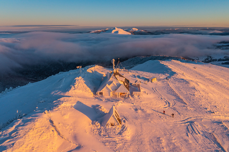 Zimowe zdjęcie z lotu ptaka. W centralnym miejscu ośnieżony budynek schronu turystycznego na szczycie góry.