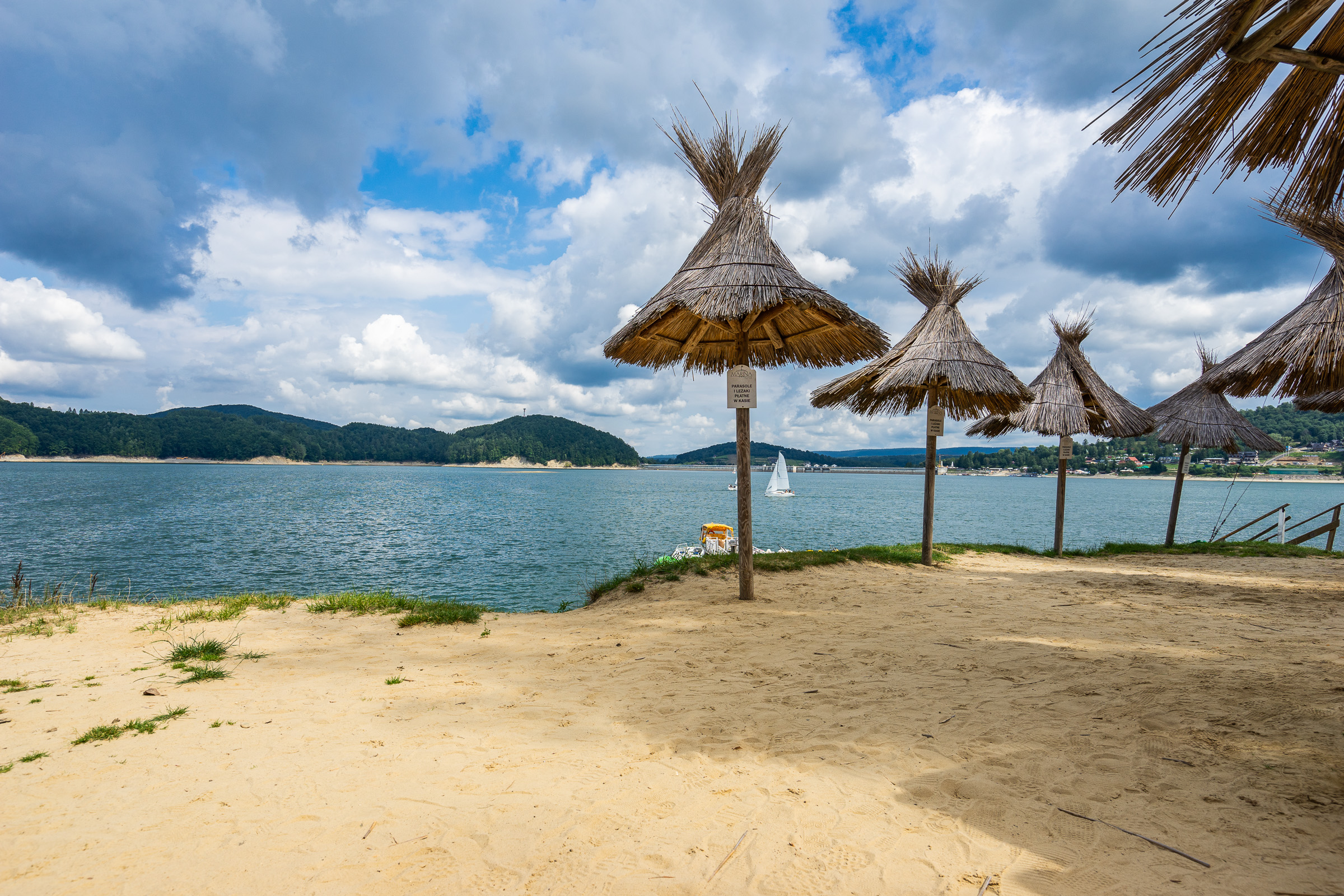Piaszczysta plaża nad jeziorem. Na plaży ustawione drewniane parasole. W tle wzgórza porośnięte drzewami i betonowa zapora. Na jeziorze kilka białych żaglówek.