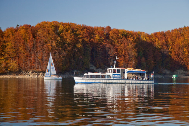 Biały statek z napisem "BRYZA" płynie po jeziorze, obok żaglówka. W tle drzewa z liśćmi w jesiennych barwach. Dominują kolory: rudy, brązowy i żółty.