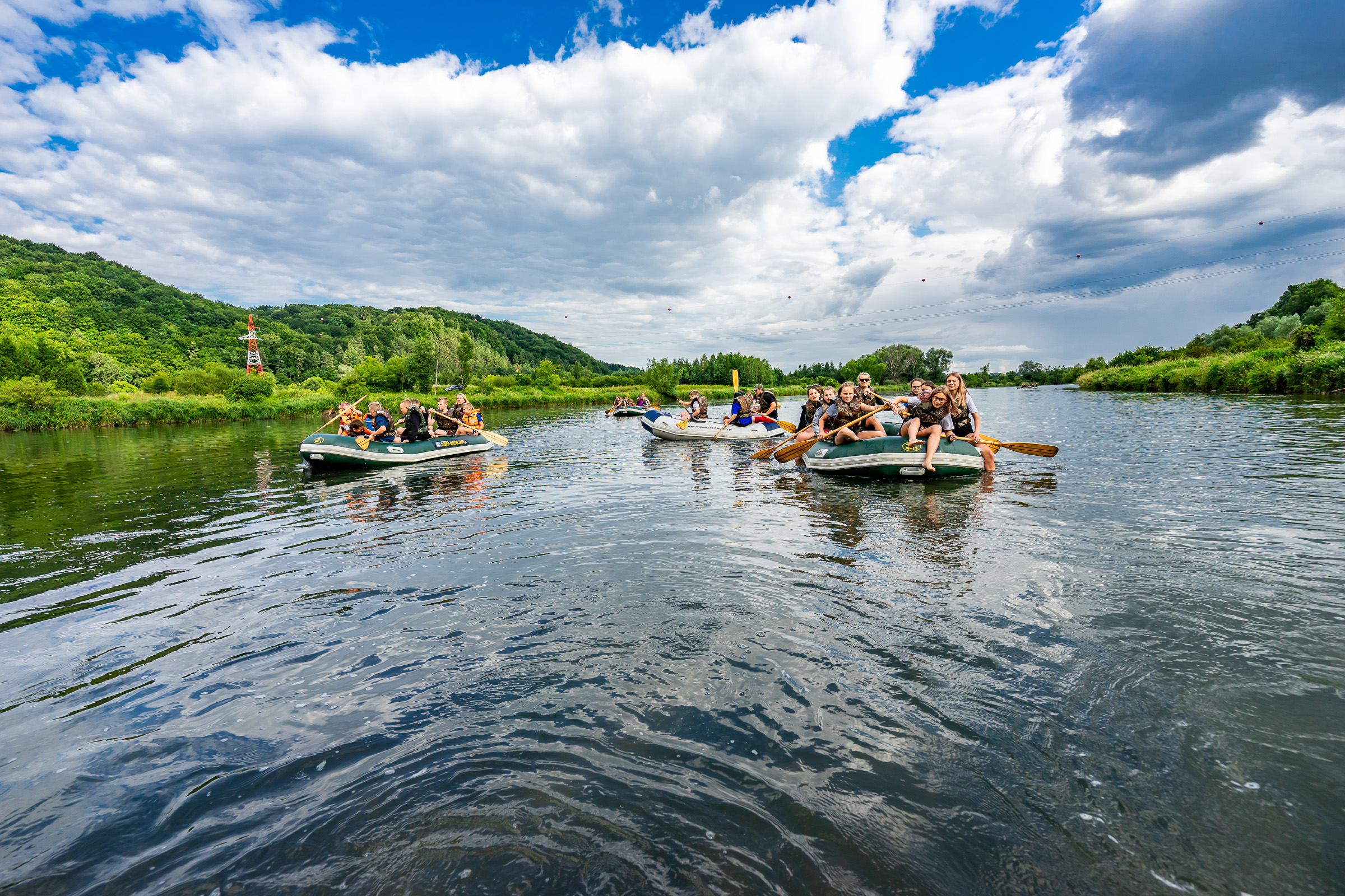 Grupa osób na kilku pontonach płynie środkiem szerokiej rzeki.
