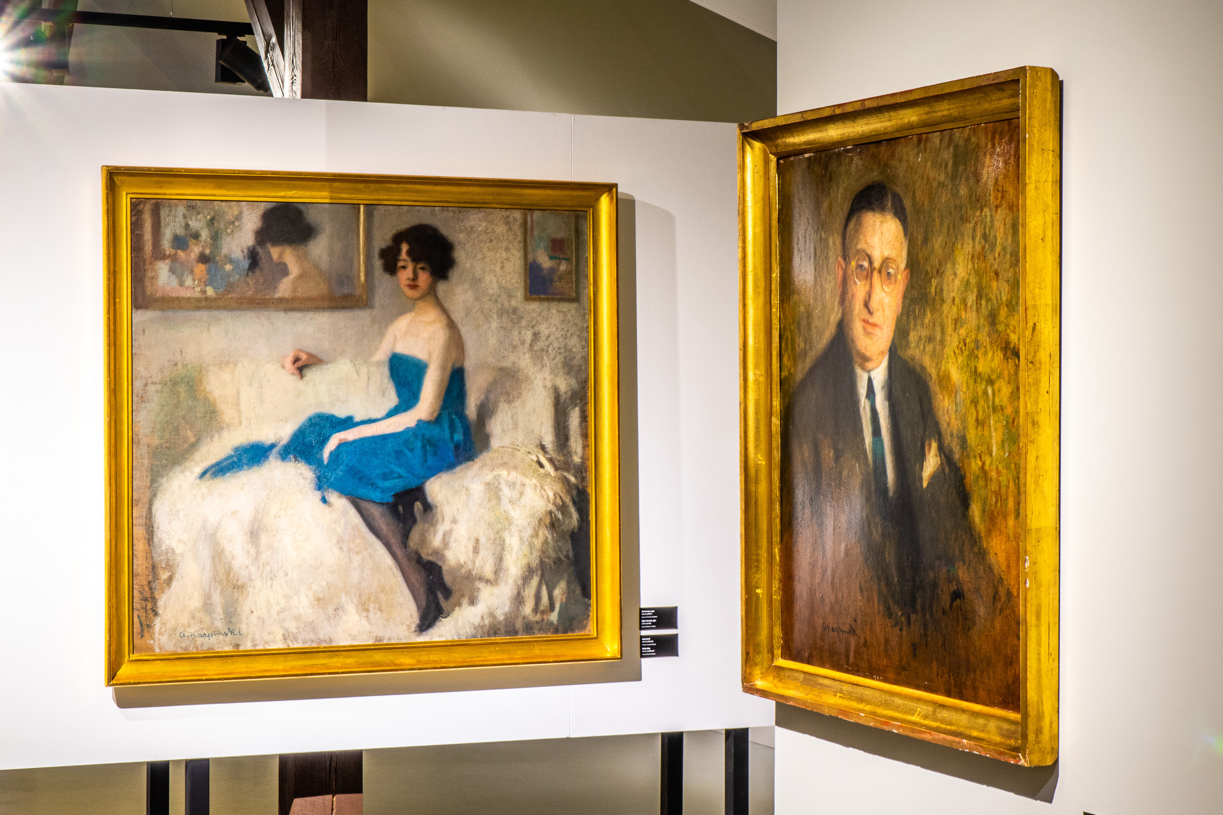 Dwa obrazy powieszone na ścianie. Na pierwszym kobieta w niebieskiej sukni siedząca na białej sofie. Na drugim portret mężczyzny w okularach i garnniturze.