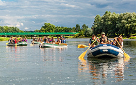 Grupa osób na kilku pontonach płynie środkiem szerokiej rzeki.