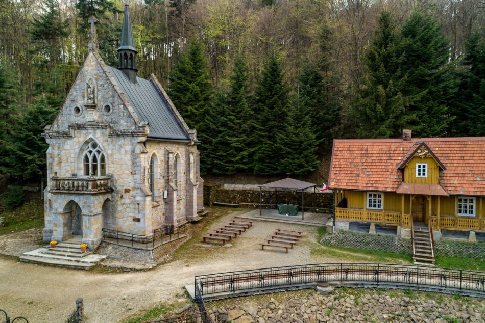 Kamienna kaplica, obok drewniany dom pośród drzew.