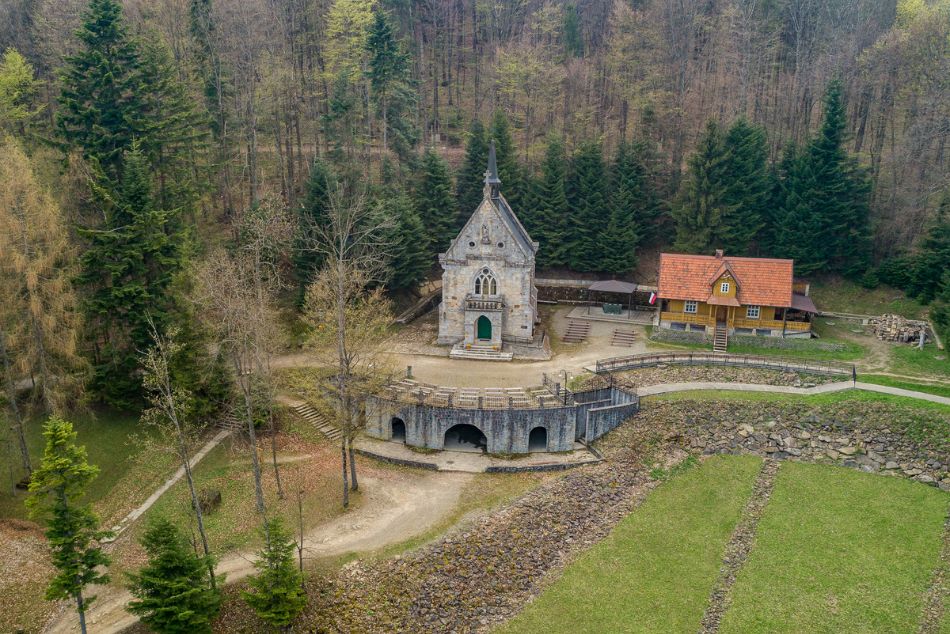 Zdjęcie z lotu ptaka. Kamienna kaplica, obok drewniany dom pośród drzew.