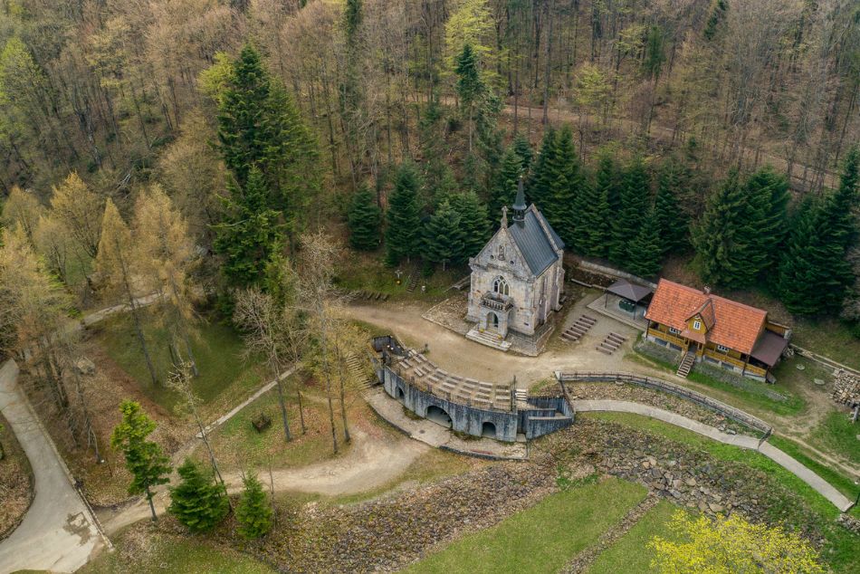 Zdjęcie z lotu ptaka. Kamienna kaplica, obok drewniany dom pośród drzew. Do kaplicczki domu prowadzi kręta droga.