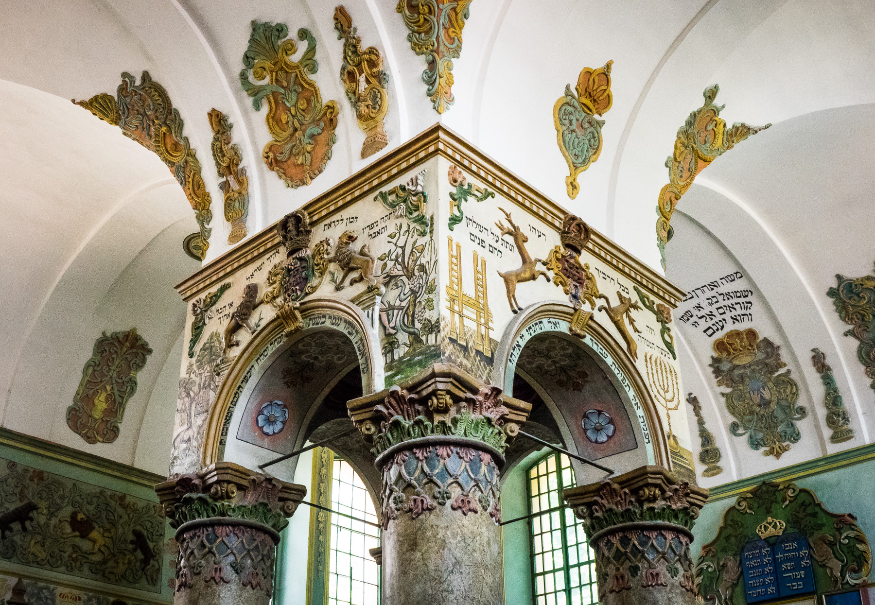 Wnętrze synagogi. W centralnym miejscu bima z bogatymi zdobieniami. Widać elementy roślinne, oraz hebrajskie napisy.