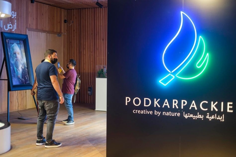 Wnętrze Pawilonu Polski na EXPO w Dubaju. Po prawej stronie na czarnym tle podświetlone niebiesko zielono logo wystawy z napisem Podkarpackie. Po lewej kilka osób zwiedzających wystawę, w tle portret Ignacego Łukasiewicza.