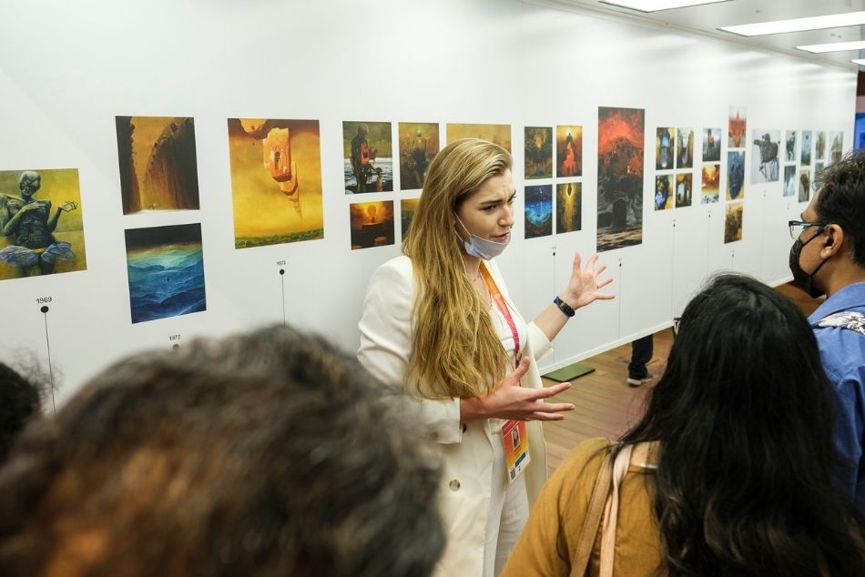 Kobieta w białym garniturze opowiada o ekspozycji obrazów Zdzisława Beksińskiego. W tle instalacja zawierająca reprodukcje najważniejszych obrazów Beksińskiego. Na pierwszym planie widać sylwetki zwiedzających.