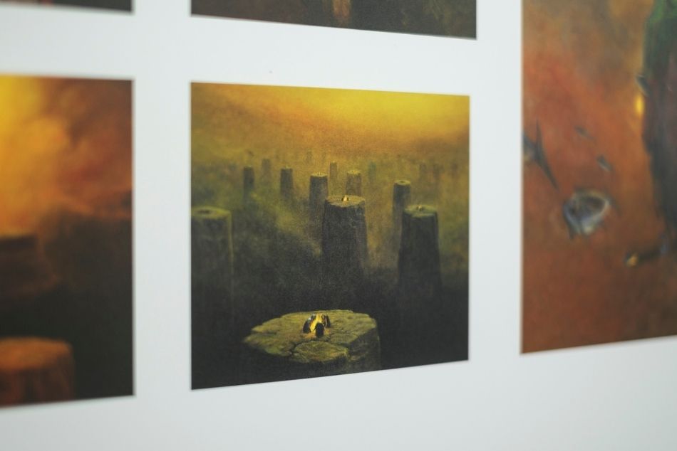 Fragment ekspozycji. Na białej planszy widać reprodukcje kilku obrazów Beksińskiego. W centralnym miejscu obraz przedstawiający kamienne obeliski spowite mgłą, na których płoną ogniska wokół których kucają postacie. Obraz utrzymany jest w brązowo-pomarańczowych barwach.