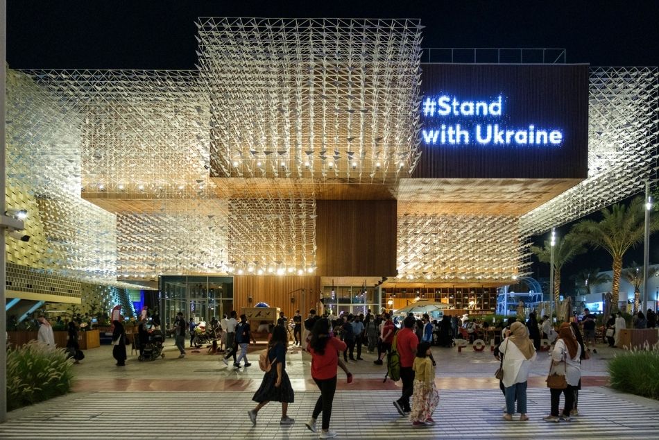 Wieczorne zdjęcie Pawilonu Polskiego na EXPO w Dubaju. Drewniany, kilkupiętrowy budynek, o nowoczesnej, geometrycznej bryle pokryty ażurową rzeźbą kinetyczną symbolizującą ptaki w locie. Na wielkim ekranie wyświetla się napis: #Stand with Ukraine