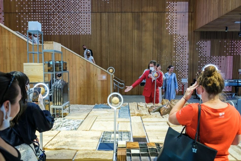 Wnętrze Pawilonu Polski. Mężczyzna w czerwonym garniturze prezentuje element drewnianej ekspozycji. Wokół kilka słuchających osób.