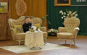 Chłopiec ucharakteryzowany na Ignacego Łukasiewicza siedzi na sofie, obok fotel, a przed nim stół
