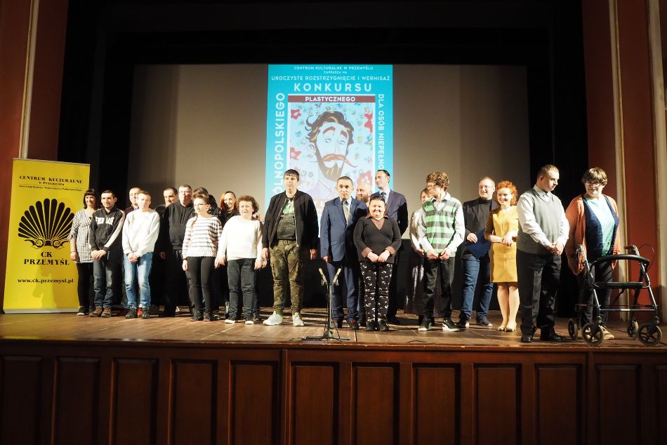 Grupa niepełnosprawnych osób na scenie. W tle plakat z wizerunkiem Ignacego Łukasiewicza oraz napisem Ogólnopolski Konkurs dla osób niepełnosprawnych.
