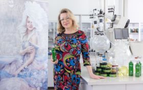 Kobieta we wzorzystej, kolorowej sukience stoi w laboratorium. Obok urządzenia laboratoryjne, oraz blat na krórym znajdują się produkowane tam kosmetyki
