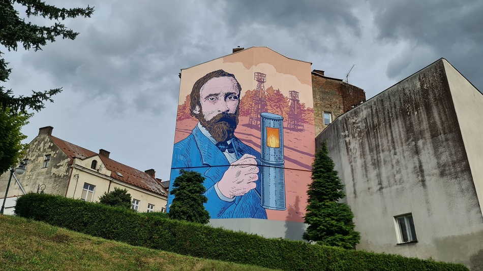 Mural z wizerunkiem Ignacego Łukasiewicza na ścianie kamienicy. Mężczyzna z brodą w niebieskim garniturze trzyma w ręku lampę naftową.