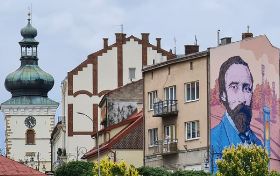 Mural z wizerunkiem Ignacego Łukasiewicza na ścianie kamienicy. Mężczyzna z brodą w niebieskim garniturze trzyma w ręku lampę naftową. Obok wieża kościelna i kamienice.