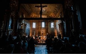Wnętrze drewnianej cerkwi. W pomieszczeniu panuje mrok, przed ołtarzem stoją muzycy.