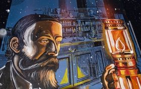 Ściana budynku z namalowanym muralem. Na pierwszym planie twarz mężczyzny z brodą i wąsami. Mężczyzna w dłoni trzyma lampę naftową. W tle laboratorium.