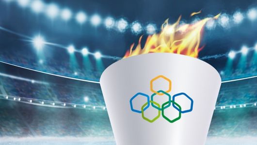 Grafika biały znicz olimpijski z płomieniem na stadionie. Na zniczu logo olimpiady młodzieżowej składające się z 6 połączonych wielobarwnych sześciokątów.