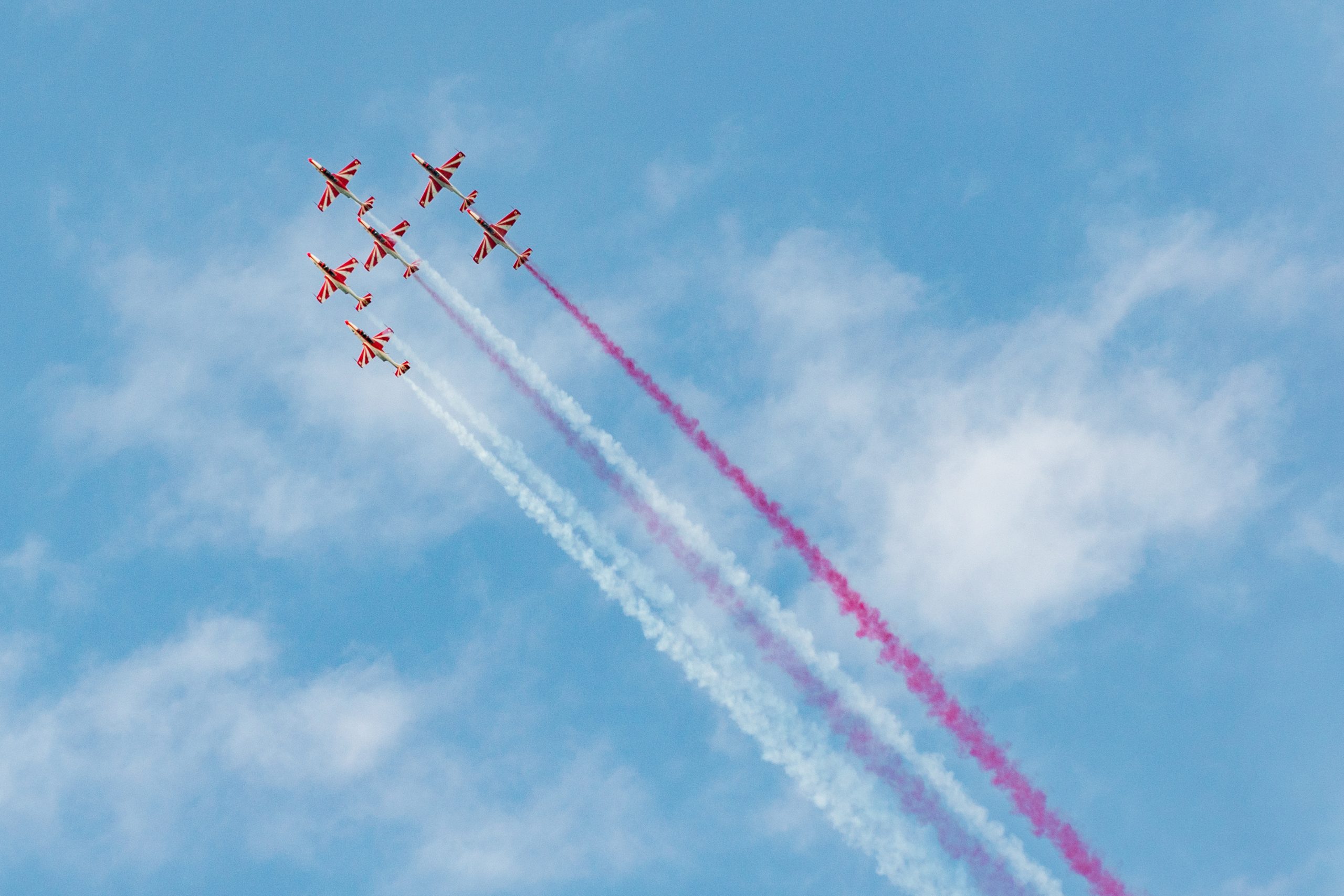 6 biało-czerwonych samolotów lecącą w zwartym szyku, za nimi ciągną się smugi białego i czerwonego dymu.