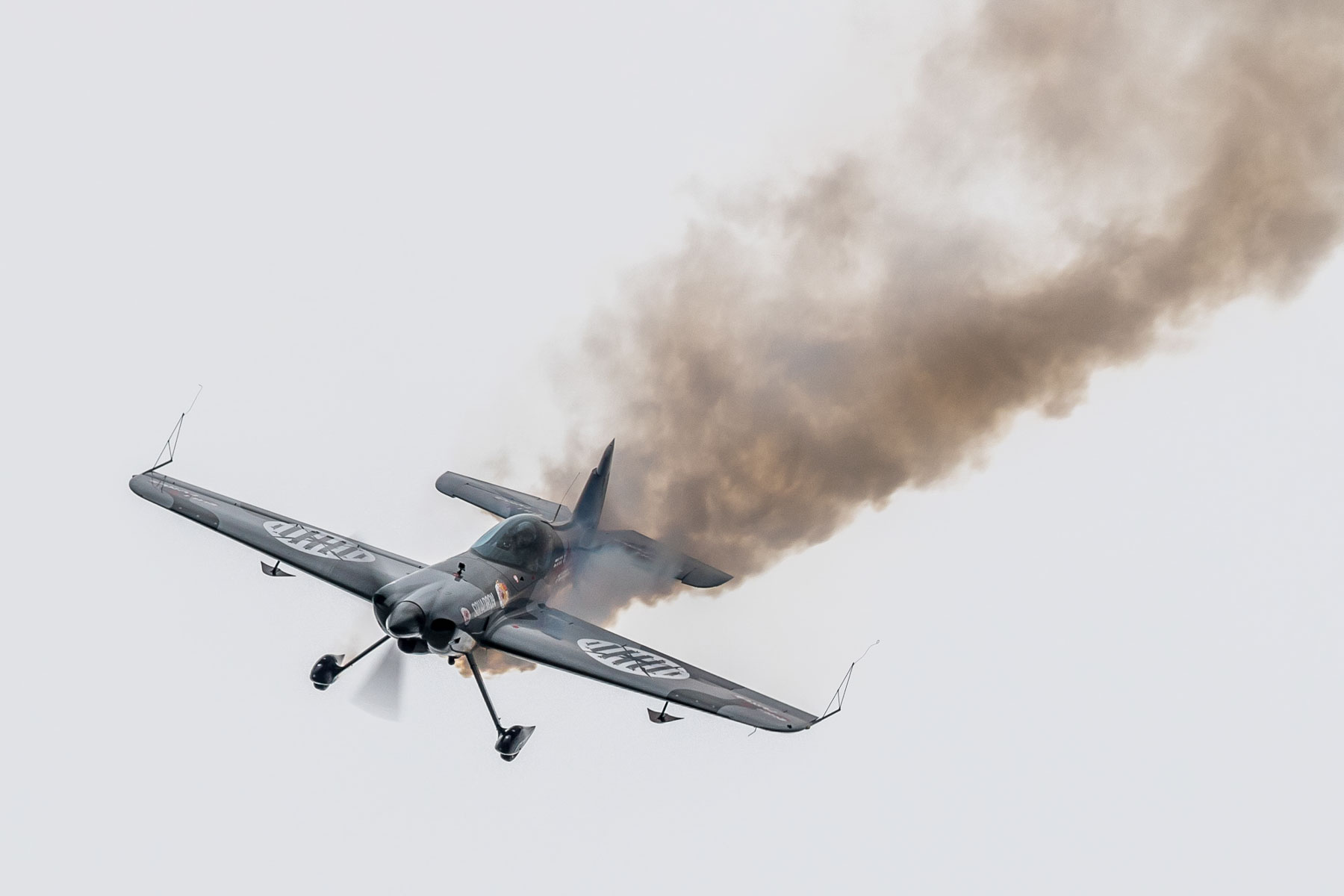 Mały samolot akrobacyjny wykonujący ewolucje na niebie. Za samolotem cięgnie się smuga dymu.