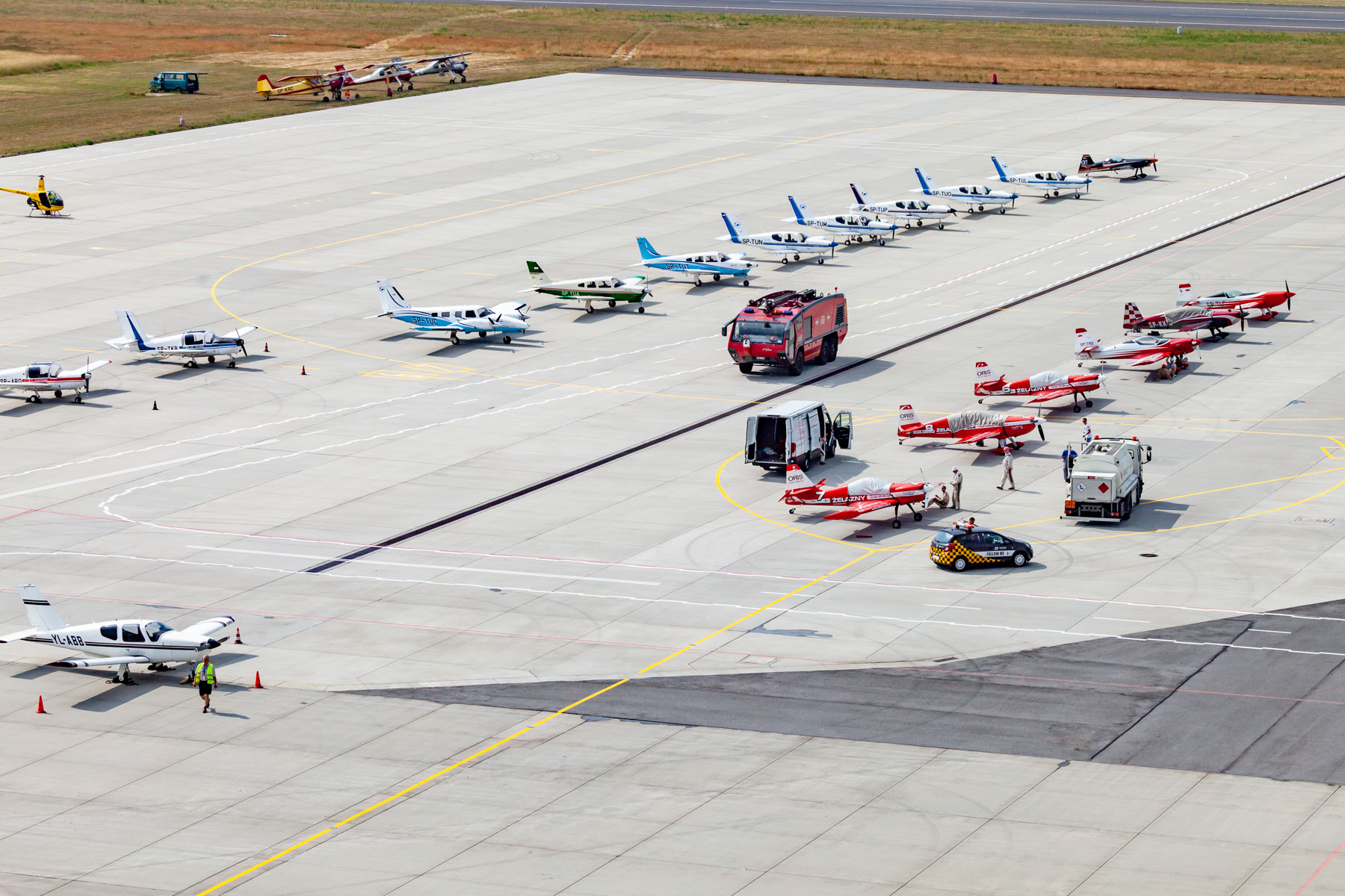 Kilkanaście samolotów stojących na płycie lotniska w dwóch rzędach. Między nimi jedzie czerwony wóz strażacki.