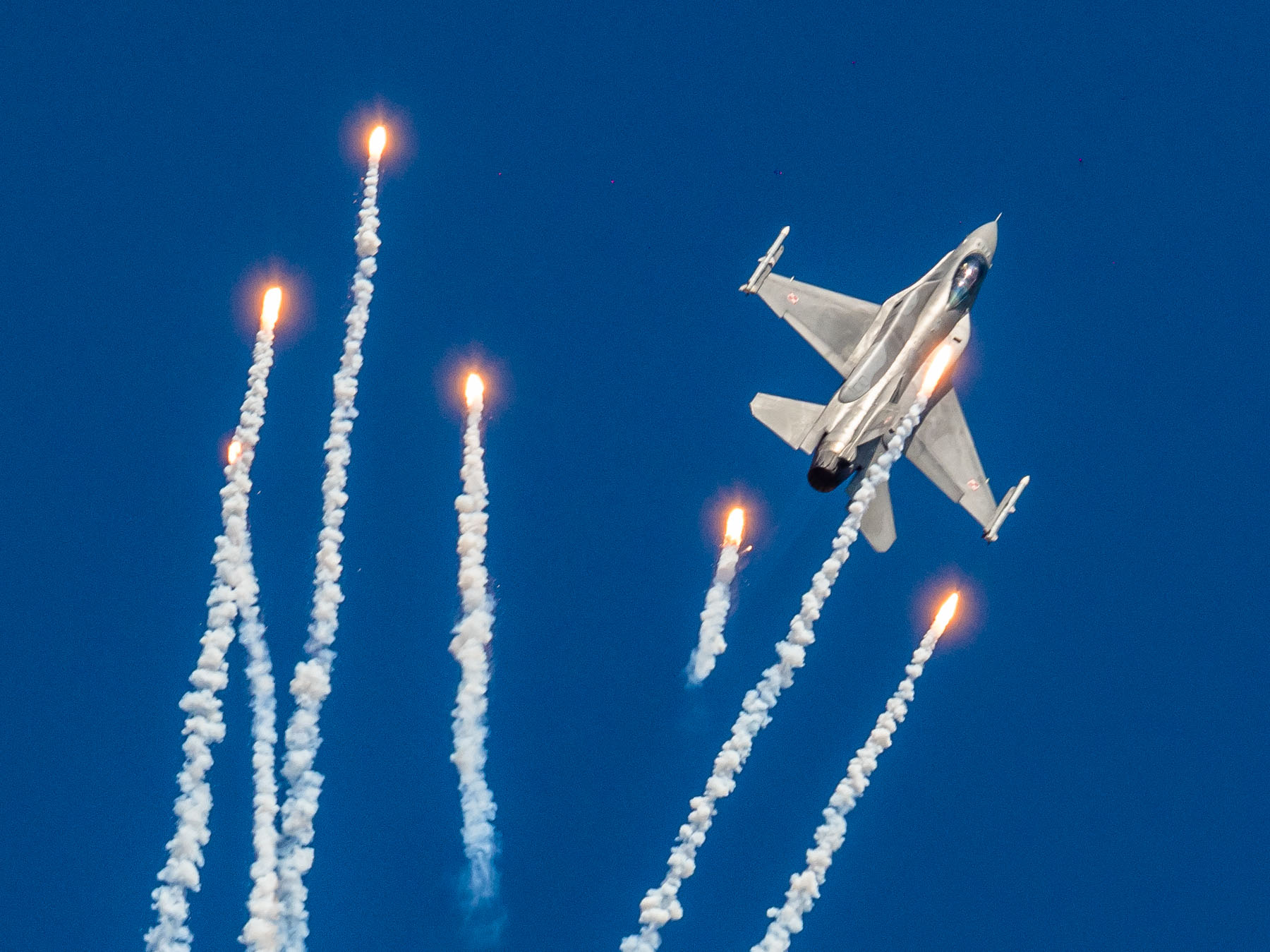 Myśliwiec wojskowy F16 leci pionowo w górę, otacza go 6 wystrzelonych flar - ognistych kul ciągnących za sobą smugę dymu.