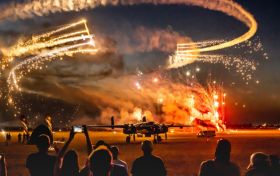 Publiczność oglądająca nocny pokaz akrobacji lotniczych połączony z pokazem pirotechnicznym. Na niebie lecą dwa samoloty za którymi ciągną się odpalone race.