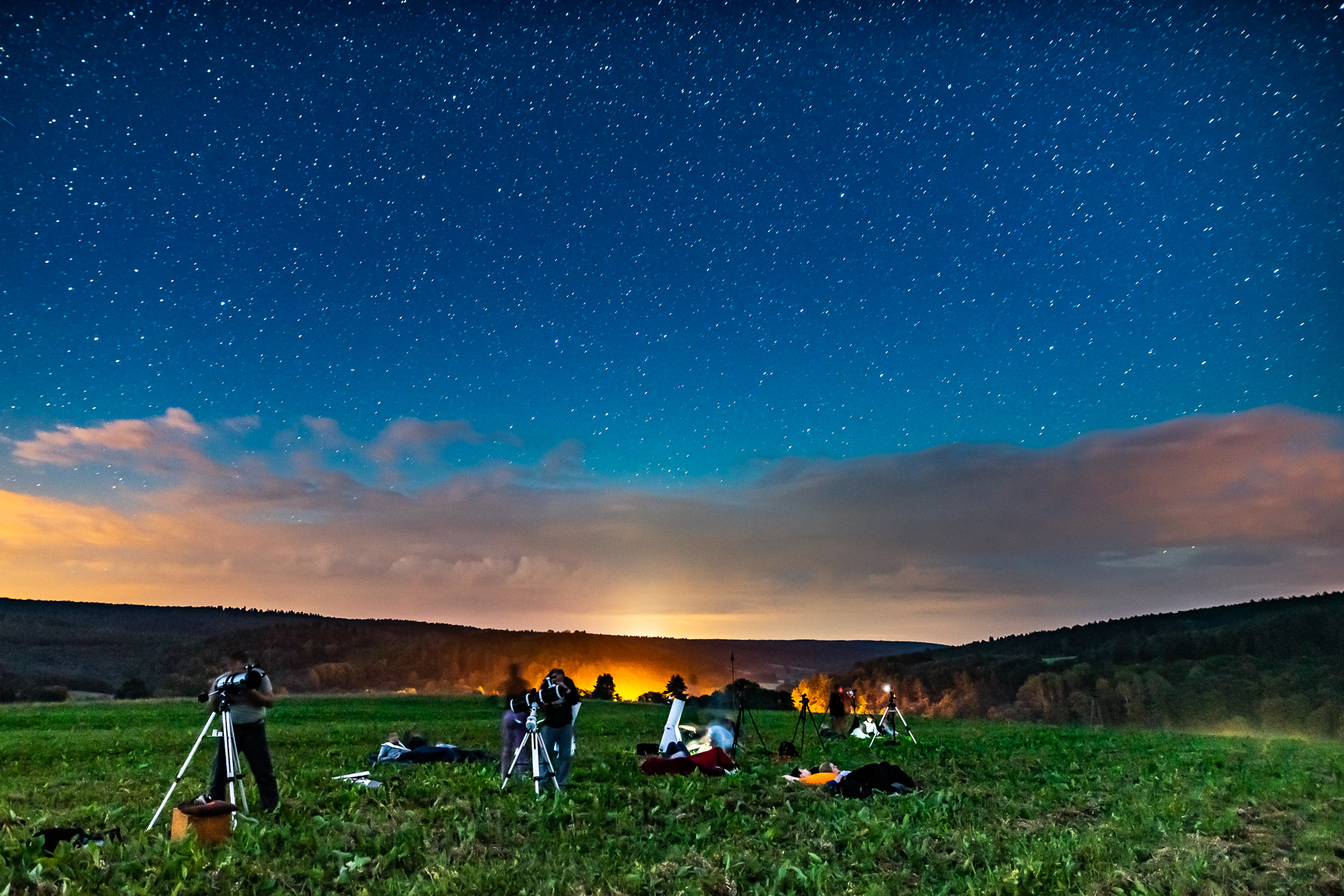 Rozgwieżdżone niebo nad górami. Na polanie grupa osób z teleskopami prowadzi obserwacje astronomiczne.