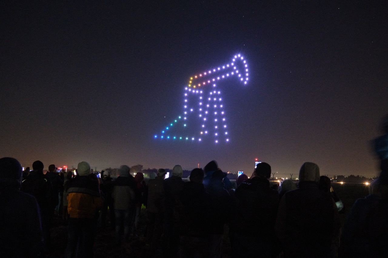 Nocny pokaz dronów. Świecące drony lecące w szyku tworzą ikonę przedstawiającą kiwon do wydobywania ropy naftowej.