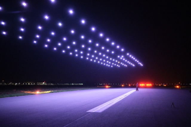 Nocny pokaz dronów. Kilkadziesiąt świecących na biało dronów lecących w szyku nad pasem startowym lotniska
