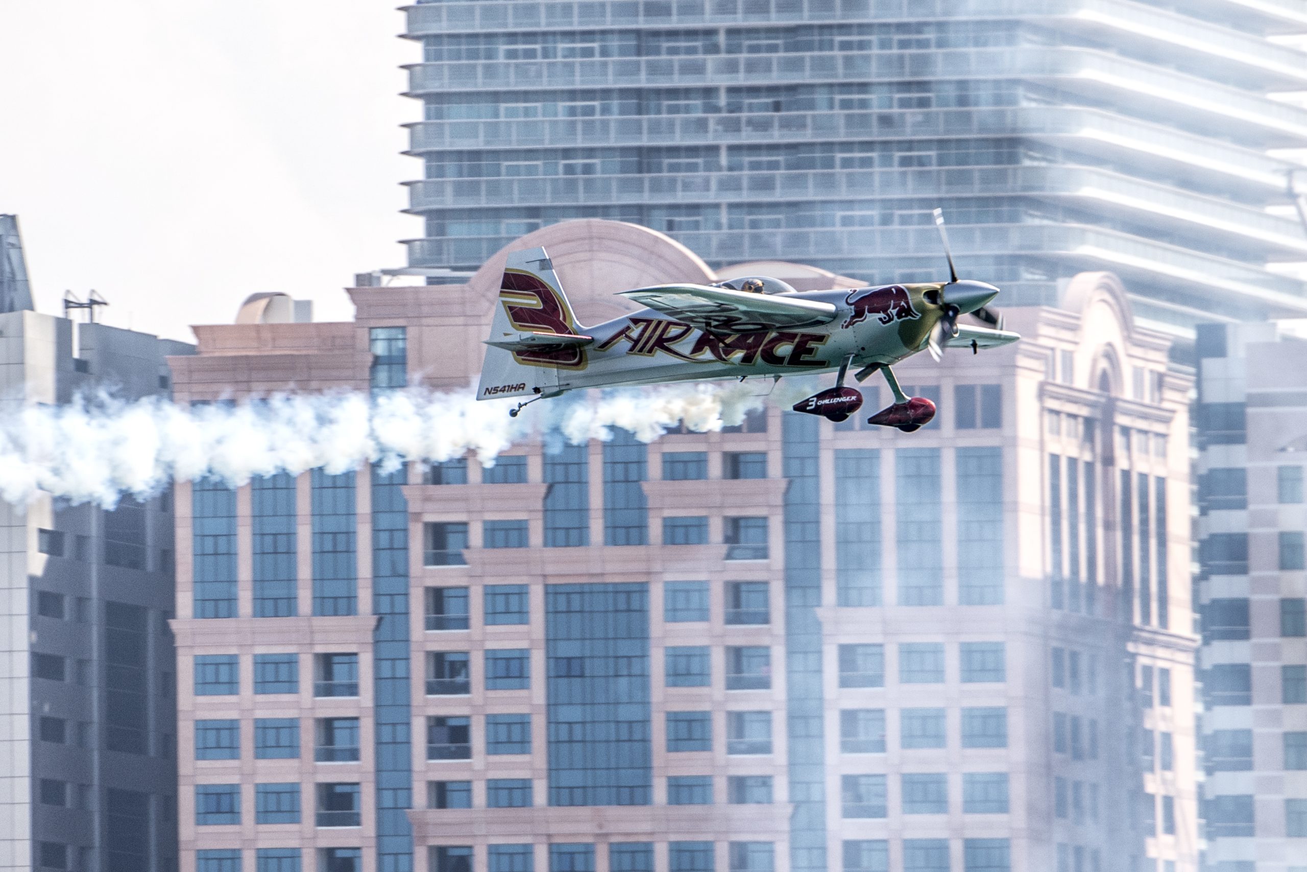 Mały samolot akrobacyjny wykonujący niski przelot na tle wysokich budynków.