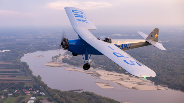 Jednopłatowy samolot z otwartą kabiną pilota leci nad rzeką.