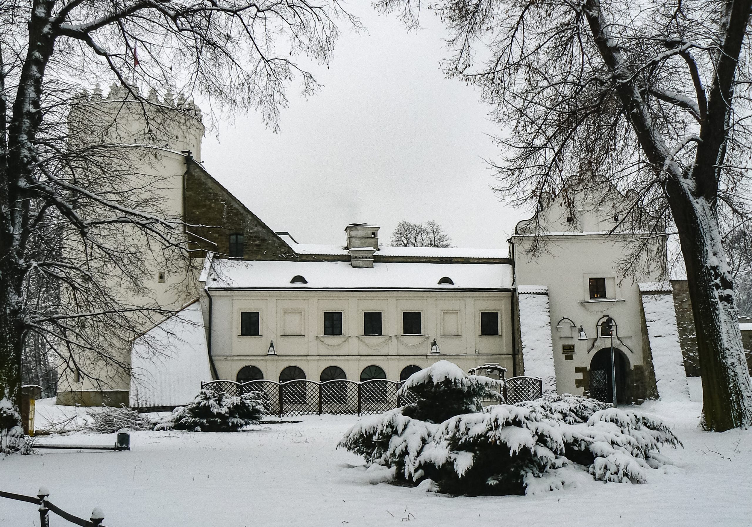 Zimowy krajobraz. Zamek z jedną wieżą i krużgankami na frontowej ścianie zewnętrznej.